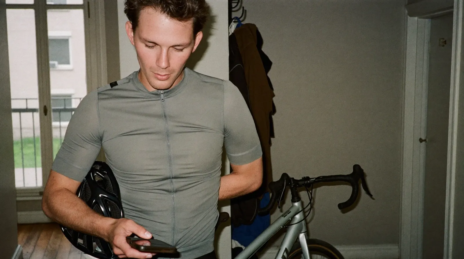Cyclist in a hallway at home, helmet under one arm, glancing down at a phone with a dark screen before heading out